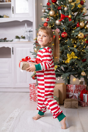 cute little child girl in Christmas pajamas with gift boxes under Christmas tree at home.の写真素材
