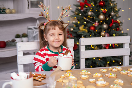 cute little girl in red Christmas pajama holding cup of cacao with marshmallows sitting on kitchenの写真素材