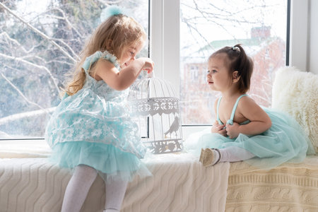 Two kids girls in blue dress sitting on a windowsill and playingの写真素材