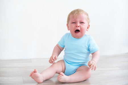 blonde baby in blue shirt crying sitting on the floor at home.の写真素材