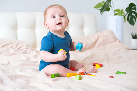 baby boy in blue bodysuit playing with colorful wooden eco toys on bed.の写真素材