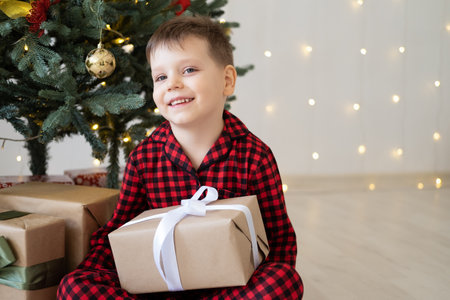 cute little child boy in red pajama with gift boxes sitting under christmas tree at homeの写真素材
