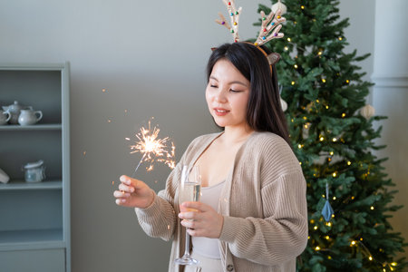 happy asian girl in cozy clothing holding glass of champagne and sparkler on her hands at homeの写真素材