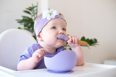 baby girl holding spoon eating pieces of fruit, sitting on high chair. first baby foodの写真素材