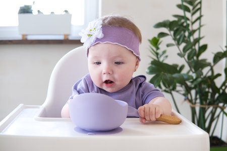 baby girl holding spoon eating pieces of fruit, sitting on high chair. first baby foodの写真素材