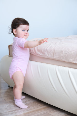 Cute baby girl in bodysuit stand holding bed frame, learning to walk.の写真素材