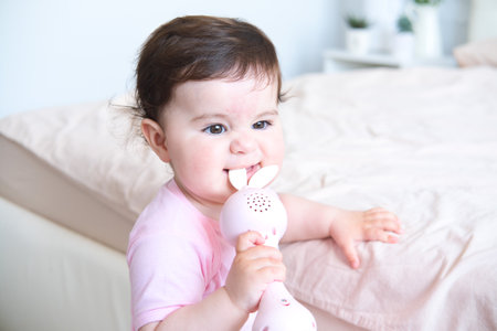 cute smiling baby with plastic bunny toy standing holding on to the bed frameの写真素材