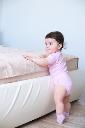 Cute baby girl in bodysuit stand holding bed frame, learning to walkの写真素材