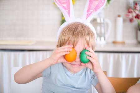 funny kid boy with bunny ears on head covering eyes with multicolored Easter eggs at home.