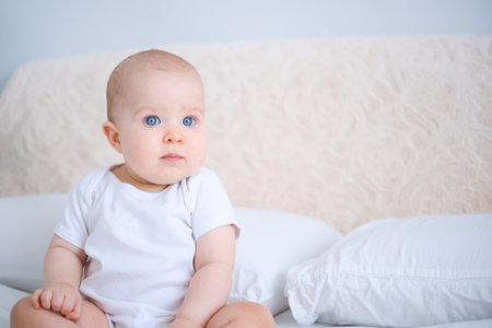 portrait of smiling healthy baby in white bodysuit sitting on bed. copy space