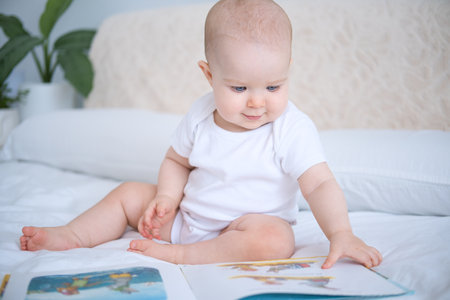 Happy funny bald baby in white bodysuit reading book sitting on parents bedの写真素材