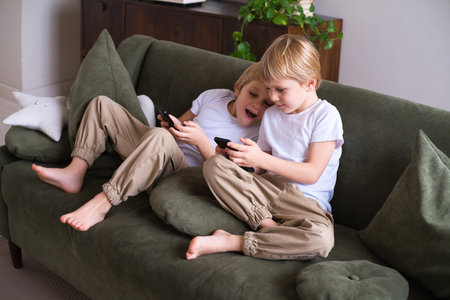 kids blonde boys twins using smartphones on the couch in living roomの写真素材