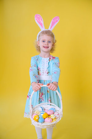 kid girl with a basket of Easter eggs and rabbit ears headband on a yellow background