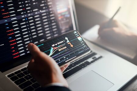 stockbroker using phone and laptop for profit gain analysis workplace. Man holding phone with bitcoin graph price.Workplace of stock trader using multiple devices for stock market analysis.の写真素材