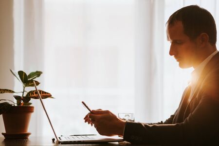 office worker using phone and laptop working day indoors. businessman using phone and laptop in office workspace with plant and coffee background. businessman using multiple gadgets.の写真素材