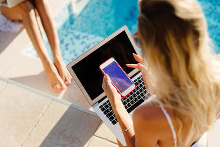 woman using laptop and smartphone remotely near swimming poolの写真素材