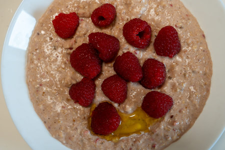 A bowl of warm oatmeal porridge topped with fresh red raspberries and a swirl of honey.