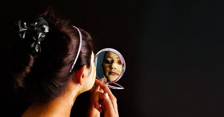 an indian lady with face pack applied in half dried condition checking her face in a mirror with selective focus on the mirror imageの写真素材