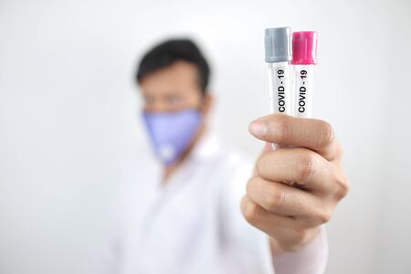 a medical professional in white coat and protective mask holding covid 19 sample vials in hand with...