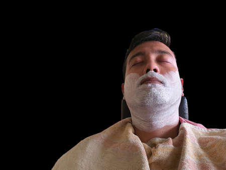 an indian man in pink shirt wearing yellow towel sitting on a chair with eyes closed and foam...