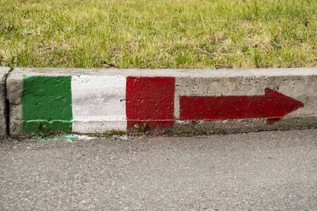 Flag of Italy with an arrow pointing the way to the right is drawn with paint on the curb on the background of asphalt and flowerbed with green grassの写真素材