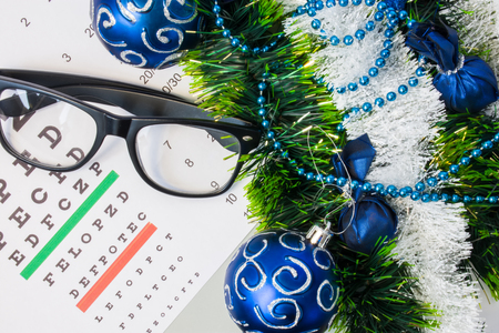 Ophthalmology or optometry Christmas and New Year with decorations. Eyeglasses and table for measure visual acuity lying near artificial snow with glitter, toys and blue balls on Christmas treeの写真素材