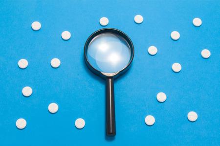 Black magnifying glass is on blue background surrounded by white pills as ornament polka dots. Medical or pharmaceutical concept by pharmacological drugs quality control, identification of drugs,pillsの写真素材