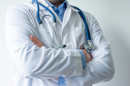 Photo of body of doctor in white lab coat with stethoscope on his neck in half-turn with folded hands against white wall in hospital with light source on left. Concept image of  medical workerの写真素材