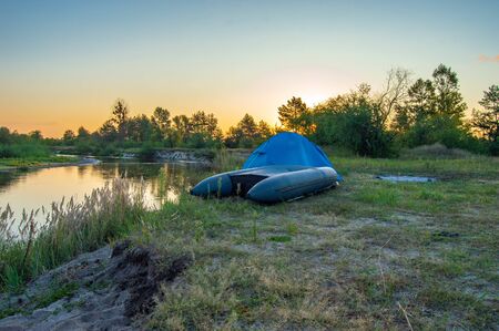 Inverted inflatable boat, pulled out of water to shore, lies near blue tourist tent on  bank of the against background of rising sun over forest. Early morning of tourist, fisherman, travelerの写真素材