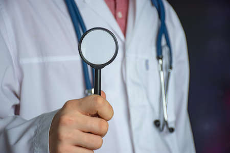 Doctor with a magnifying glass in his hands against the background of a white coat. Concept photo for diagnosing and searching for diseasesの写真素材