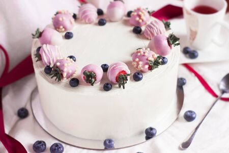 Festive white cake with strawberries in chocolate and fresh blueberries on a white background. Side...