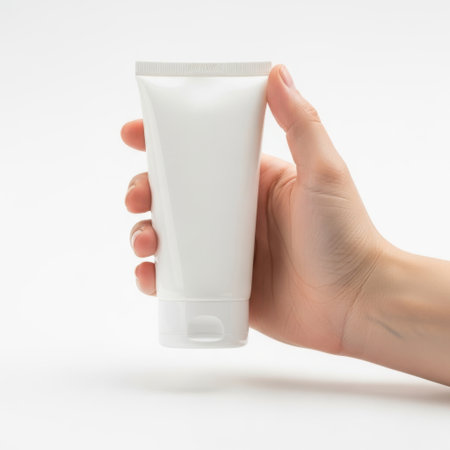 The image shows a hand holding a tube of cream against a plain white background. The tube is cylindrical with a flip cap and appears to contain a white cream or lotion. The hand is positioned with the fingers supporting the tube from the bottom and the thumb on the side.の素材