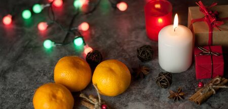 Festive background on which a white candle burns on a table among gifts in craft paper, rattan balls, spruce wreath, tangerines, cinnamon sticks on the background of a multi-colored Christmas garland and red tinsel with snowflakesの写真素材