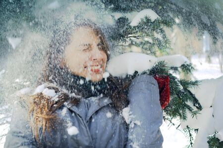 Young Ukrainian girl standing under spruce branches covered in snow in the park. The girls are wearing winter closes and squints in joy when the snow is falling on her from the treeの写真素材