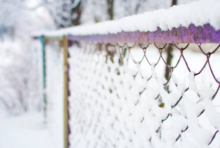 Closeup of a purple mesh fence covered in a thick layer of white fresh fluffy snow against blurred trees on the background. Cold winter day in Januaryの写真素材
