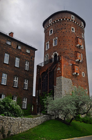 One of the high towers of the Wawel castle in Krakow with loopholes surrounded by fresh green grass, flowering trees and bushes. Dark medieval atmosphere on a cloudy day in springの写真素材