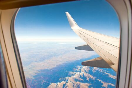 White airplane wing shor from a window. Aerial view of majestic mountain range with snow-capped peaks, green and red forest, fields, blue sky and horizon. Sunny cloudless autumn dayの写真素材