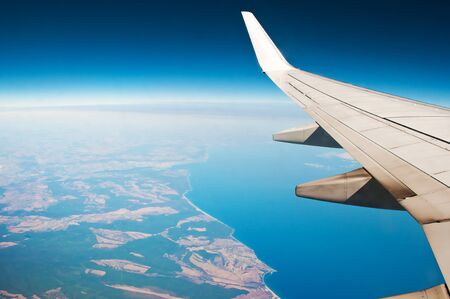 White airplane wing. Aerial view of blue sea surface, ragged sea coast, fields, sky and horizon. Air travel on a sunny cloudless dayの写真素材