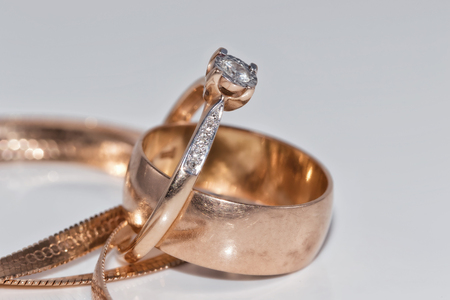 Thin elegant gold diamond ring, thick wedding ring and a gold chain with a snake tribe on a white reflective surfaceの写真素材