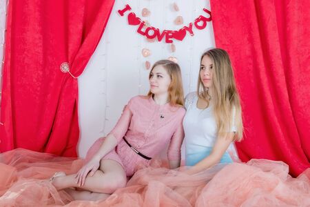 Two cute long-haired girlfriends in dresses sit and cuddle against the background of red velvet curtainsの写真素材