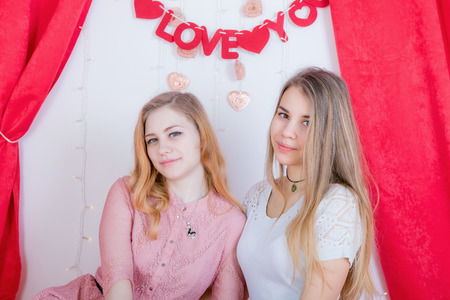 Two cute long-haired girlfriends in dresses sit and cuddle against the background of red velvet curtainsの写真素材