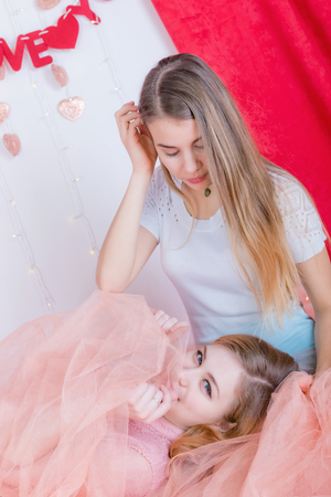 Two cute long-haired girlfriends in dresses sit and cuddle against the background of red velvet curtainsの写真素材