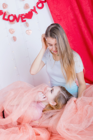 Two cute long-haired girlfriends in dresses sit and cuddle against the background of red velvet curtainsの写真素材