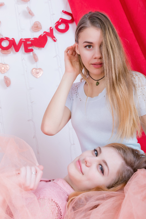 Two cute long-haired girlfriends in dresses sit and cuddle against the background of red velvet curtainsの写真素材