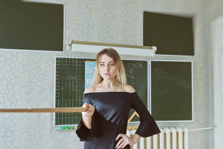 Young slender beautiful blonde teacher in a black dress with a pointer stands at the blackboard in the classroomの写真素材