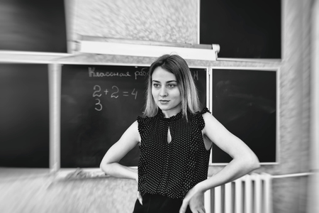 Strict young teacher blonde standing resting her hands on her hips at the blackboard in class and looking at the audienceの写真素材