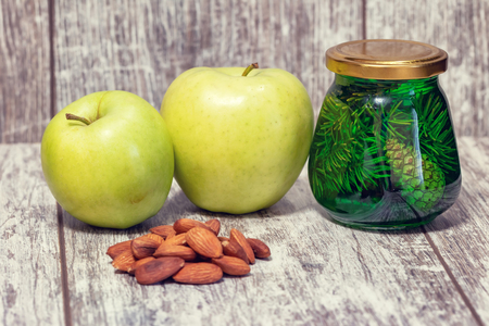 Two large green apples, a handful of almonds and a jar of jam from fir cones are on the table top of white oakの写真素材