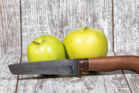 Two large green apples and a knife with a blade of Damascus steel lie on the table top of white oakの写真素材
