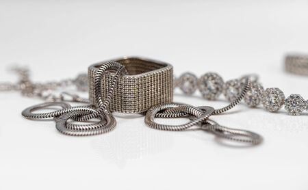 Square silver diamond ring and silver snake chain on white reflective surfaceの写真素材
