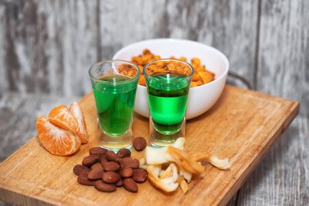 Two shot of absinthe stand on a wooden board. Next to it is an appetizer : fried almonds, smoked cheese and Mandarin slicesの写真素材